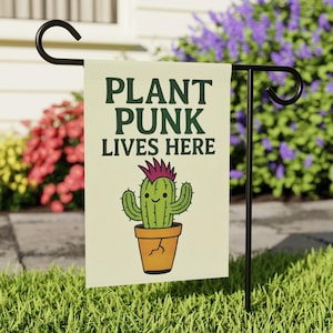 Puede incluir: Una bandera de jardín con el texto "PLANT PUNK LIVES HERE" en verde oscuro. La bandera presenta un cactus de dibujos animados con una cresta rosa en una maceta naranja. La bandera se muestra en un soporte de metal negro en un jardín.