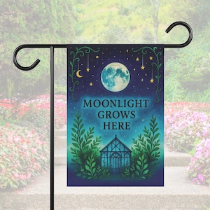 以下が含まれることがあります： 夜空のデザインのガーデンフラッグ。旗には満月、星、そして「MOONLIGHT GROWS HERE」というテキストが描かれています。緑の葉と温室も描かれています。旗は黒い金属製のスタンドに表示されています。