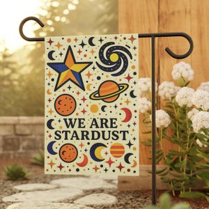 May include: A rectangular garden flag with a black metal stand. The flag features a colorful celestial design with a star, planets, moons, and the text "WE ARE STARDUST". The flag is cream-colored with a variety of colors.