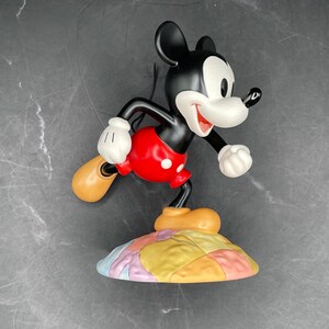 May include: A figurine of Mickey Mouse in a running pose. The figure is black, white, and red, with tan shoes and white gloves. Mickey is standing on a colorful, patchwork-style base. The figurine is a collectible item.