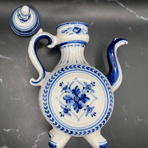 Puede incluir: Jarra de cerámica blanca y azul con tapa a juego. La jarra presenta un diseño floral en azul y blanco, con un asa y un pico curvos. La jarra está decorada con motivos florales azules y un borde decorativo.