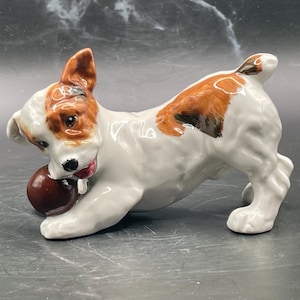 May include: A white and brown porcelain figurine of a dog playing with a brown ball. The dog has brown spots on its back and a pink tongue sticking out.