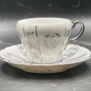 May include: A white teacup and saucer with silver floral designs. The cup has a silver handle and the saucer has a scalloped edge.