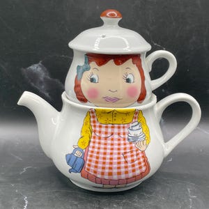 May include: White ceramic teapot and teacup set featuring a cartoon girl illustration. The teapot has a spout and handle, and the teacup sits on top. The girl wears a yellow dress and a red and white checked apron.