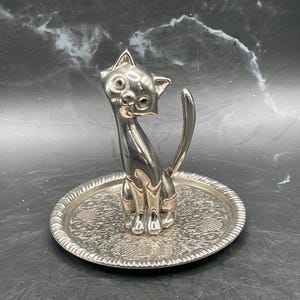 May include: A silver cat-shaped ring holder. The cat sits on a round, patterned tray. The cat has large eyes and a curved tail. The ring holder is made of metal and has a shiny finish. The tray has a diameter of approximately 10 cm.
