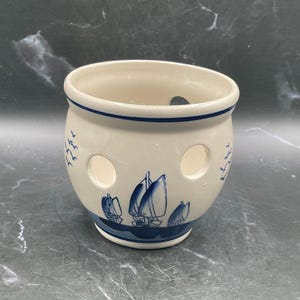 May include: A white ceramic pot with a blue rim and a blue sailboat design. The pot has three circular holes and blue bird silhouettes. The pot is a decorative item.