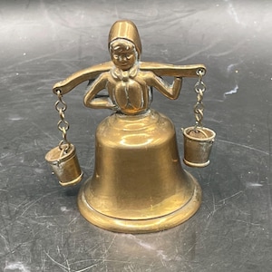 May include: A vintage brass bell shaped like a woman carrying buckets. The bell has a detailed figure of a woman wearing a bonnet and holding a yoke with two small buckets suspended from chains. The bell is a warm gold colour.