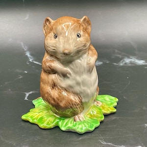 May include: A brown and white ceramic figurine of a mouse sitting on a green leaf. The mouse has a brown body, white belly, and black eyes.