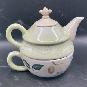 Peut inclure: Service à thé individuel en céramique aux couleurs pastel. La théière a un couvercle et un corps vert clair, avec un bouton et un bec verseur crème. La tasse est décorée de motifs floraux et de feuilles.