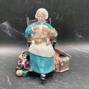 May include: A ceramic figurine depicting a woman in a blue dress and white apron, seated in a brown chair, holding a teddy bear. A small doll and a basket are at her feet. The figurine has a vintage appearance.
