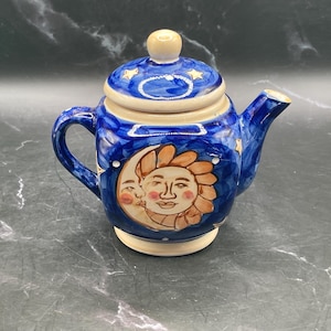 May include: A ceramic teapot in blue and cream. The teapot has a lid with a small knob and is decorated with a sun and moon design. It has a handle and a spout. The teapot is suitable for serving tea.