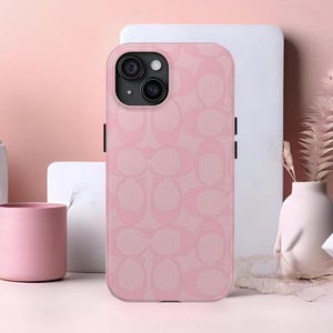 May include: Pink phone case with a repeating pattern. The case has a camera cutout and black side buttons. The background is a soft pink with a white rectangular shape and a pink vase.