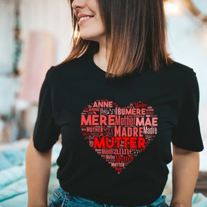 Puede incluir: Una camiseta negra con un diseño en forma de corazón rojo. El corazón está formado por la palabra "madre" en varios idiomas, como "Mutter", "Madre", "Mere" y "Mother".