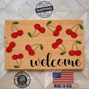 May include: Rectangular doormat with a tan background, featuring a pattern of red cherries with green stems. The word "welcome" is written in black script. Includes a "Made in USA" flag.