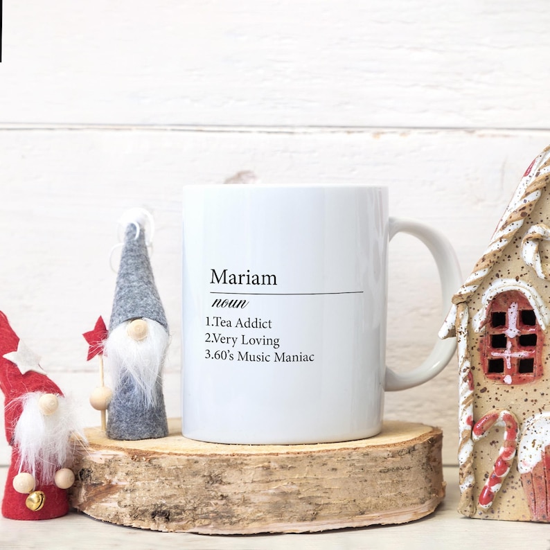 May include: White ceramic mug with a black design that says "Mariam noun 1.Tea Addict 2.Very Loving 3.60's Music Maniac"
