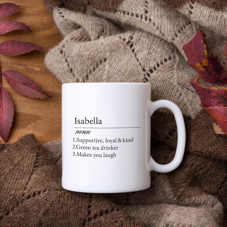 May include: White ceramic mug with a black inscription that reads "Isabella noun 1.Supportive, loyal & kind 2.Green tea drinker 3.Makes you laugh" The mug is sitting on a brown wooden surface with a beige knitted blanket and red leaves.