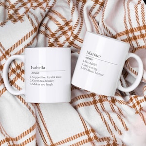 May include: Two white ceramic mugs with black text. The mug on the left says "Isabella noun 1.Supportive, loyal & kind 2.Green tea drinker 3.Makes you laugh". The mug on the right says "Mariam noun 1.Tea Addict 2.Very Loving 3.60's Music Maniac".