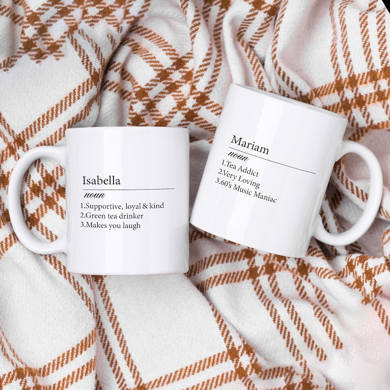 May include: Two white ceramic mugs with black text. The mug on the left says "Isabella noun 1.Supportive, loyal & kind 2.Green tea drinker 3.Makes you laugh". The mug on the right says "Mariam noun 1.Tea Addict 2.Very Loving 3.60's Music Maniac".