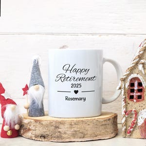 Pu&ograve; includere: Una tazza in ceramica bianca con testo nero che recita "Happy Retirement 2025" con un simbolo di cuore e il nome "Rosemary" sotto. La tazza &egrave; appoggiata su una superficie di legno con una piccola statuetta di gnomo a sinistra.