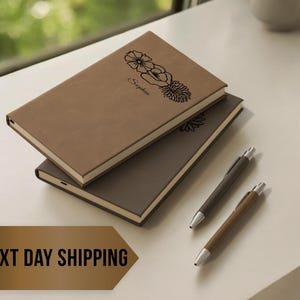 May include: Two brown leather-bound journals with floral designs and the word "Simple" on the top journal. Two pens are also included.