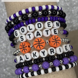 May include: A stack of colorful beaded bracelets with the words "GOLDEN STATE" and "ALKYRIES" spelled out in white beads. The bracelets also feature orange basketballs with black stitching.