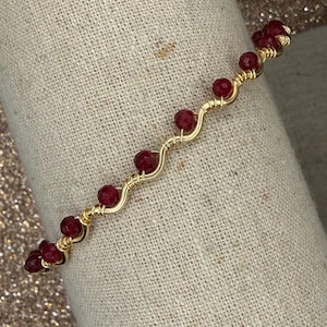 May include: A gold-toned wire bracelet with a wavy design, featuring a series of deep red beads. The beads are evenly spaced along the wire, creating a delicate and elegant accessory. The bracelet is displayed on a neutral-toned fabric.