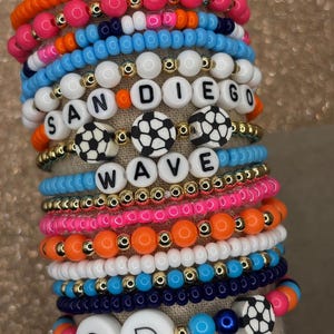 May include: A stack of colorful beaded bracelets with white letter beads spelling out "SANDIEGO WAVE" and "SD". The bracelets also feature soccer ball charms. The text "Bracelets sold individually or as a STACK!" is at the bottom of the image.