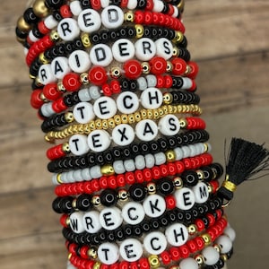 May include: Stack of beaded bracelets in red, black, white, and gold. Some bracelets have white letter beads spelling out "RED", "RAIDERS", "TECH", "TEXAS", "WRECKEM", and "TECH". A black tassel is attached.