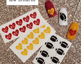 May include: Two sheets of nail stickers with a Kansas City Chiefs theme. One sheet has red hearts with white "KC" inside. The other sheet has yellow hearts with white "KC" inside. The third sheet has black footballs on a white background.