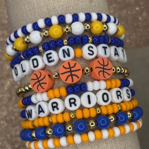 May include: A stack of beaded bracelets with the words "Golden State Warriors" spelled out in white letters. The bracelets are made with blue, white, yellow, and orange beads. The bracelets are sold individually or as a stack.
