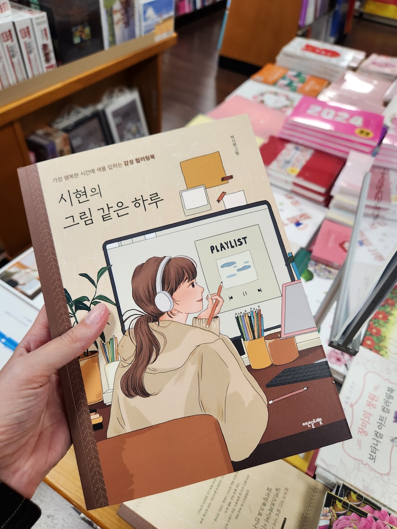 May include: A book cover with a brown background and a colourful illustration of a woman wearing headphones and drawing on a tablet. The title of the book is "시현의 그림 같은 하루" which translates to "Sihyun's Day Like a Painting".