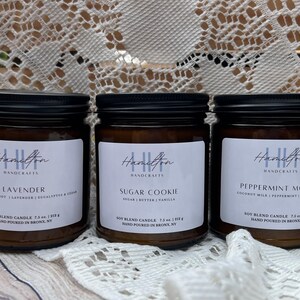 May include: Three amber glass candle jars with black lids. The labels are white with black text. The candles are labeled "Lavender", "Sugar Cookie", and "Peppermint Mocha".