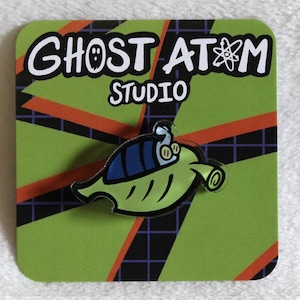 May include: A green and blue cartoon spaceship pin with a black outline. The pin is on a green and orange background with a grid pattern. The text "Ghost Atom Studio" is printed on the background.