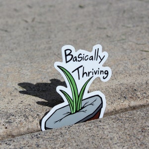 May include: A white sticker with the words "Basically Thriving" in black script. The sticker features a cartoon image of green grass growing out of a gray rock. The sticker is placed on a concrete surface.
