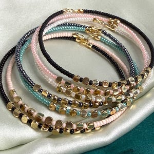 May include: A collection of five circular beaded bracelets featuring small, round, matte-finish beads in black, light pink, and teal, interspersed with metallic gold-toned flat disc beads. Each bracelet is secured with a small gold-toned metal clasp.