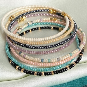 May include: A stack of seven thin beaded bracelets featuring small, uniform glass beads in various colors including cream, black, metallic grey, soft pink, lavender, and turquoise, accented with small gold-toned metal spacer beads throughout.