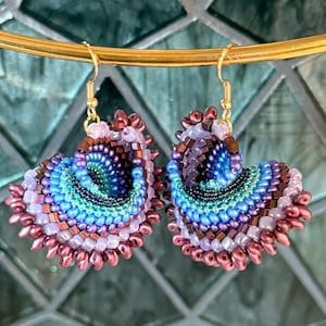 May include: A pair of dangle earrings featuring a spiral shell design crafted from multi-colored seed beads in shades of metallic bronze, lavender, bright blue, and teal. The earrings hang from gold-toned metal hooks and have a textured finish.