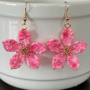 Puede incluir: Un par de pendientes colgantes con forma de flor de cinco pétalos, elaborados con pequeñas cuentas de semillas de color rosa vibrante. El centro de cada flor está acentuado con cuentas doradas metálicas y cuelgan de ganchos de metal dorado.