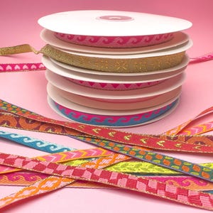 Può includere: Una pila di rocchetti di nastro decorativo in vari colori e fantasie, tra cui rosa, oro, blu e arancione. I nastri presentano disegni a onde, cuori e geometrici. Lo sfondo è rosa pieno.