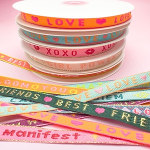 Könnte beinhalten: Eine Sammlung dekorativer Bandspulen in verschiedenen Farben und mit Texten wie "LOVE", "XOXO" und "BEST". Die oberste Spule hat ein orangefarbenes Band mit dem Wort "LOVE" und einem Herzen. Weitere Bänder sind sichtbar.
