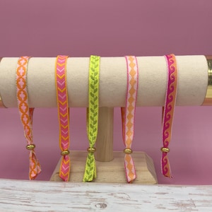 Może przedstawiać: Pięć kolorowych, tkanych bransoletek z różnymi wzorami, prezentowanych na białym stojaku. Bransoletki są różowe, pomarańczowe, zielone i żółte.