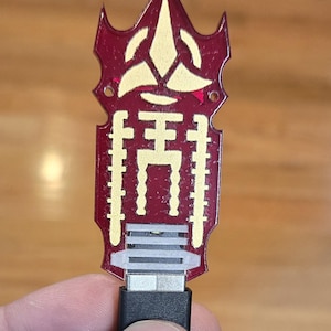 May include: A decorative USB-C charging cable with a burgundy and gold design. The top of the cable has a stylized shape with a gold emblem and a series of gold vertical lines. The cable is held by a hand.