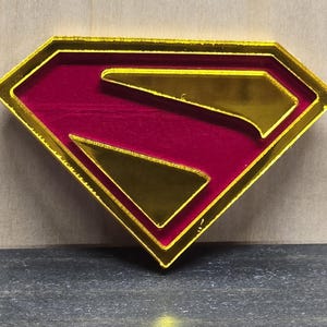 May include: A gold-edged, red and gold Superman logo. The iconic 'S' shield is a diamond shape with a gold outline and a red center. The gold 'S' is visible within the red background. The logo is set against a neutral background.