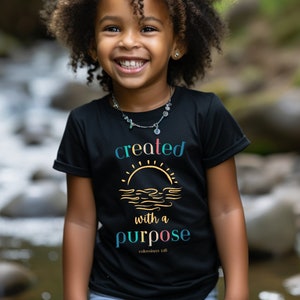 May include: A young girl wearing a black t-shirt with a gold sun and waves graphic and the text "created with a purpose" in rainbow colors. The text "colossians 1:16" is printed in smaller font below the graphic.