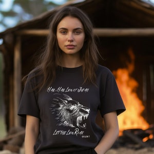 以下が含まれることがあります： ライオンの頭が吠えている白いグラフィックがプリントされた黒いTシャツ。シャツには「Hail, Hail Lion of Judah」と「Let the Lion Roar」の文字がプリントされています。シャツは、後ろに火のある木造の構造物の前に立っている人が着用しています。