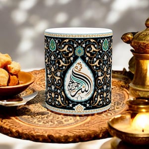 May include: A white ceramic mug with a black and gold design featuring intricate floral patterns and Arabic calligraphy. The mug is sitting on a wooden tray with a gold-coloured metal candle holder.