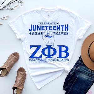 Könnte beinhalten: Weißes T-Shirt mit blauem Text und Grafiken. Das Shirt trägt "Celebrating Juneteenth" über einer erhobenen Faust-Grafik und "ZΦB" darunter. Dekorative geometrische Muster umranden den Text. Das Shirt wird mit Jeans, einem Hut und braunen Sandalen kombiniert.