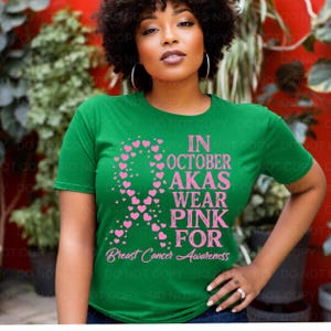 May include: A green short-sleeve t-shirt featuring a pink awareness ribbon composed of small hearts and the text IN OCTOBER AKAS WEAR PINK FOR Breast Cancer Awareness printed in pink on the front of the garment.