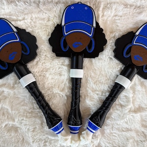 May include: Three decorative bottle stoppers featuring a stylized face with a blue baseball cap, blue lipstick, and hoop earrings. The stoppers have black handles with white and blue accents, and are set against a white, fluffy background.
