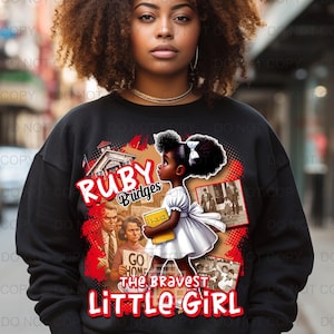 Op de afbeelding: Zwart sweatshirt met een afbeelding van Ruby Bridges, een jong zwart meisje, en de tekst "Ruby Bridges" en "The Bravest Little Girl". Het ontwerp bevat afbeeldingen van een school, demonstranten en historische foto's.